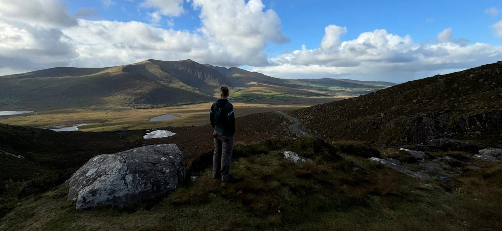 A man standing on a rocky viewpoint looking across a wide valley with small lakes to a large mountain